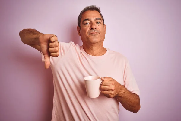 Handsome middle age man holding cup of coffee standing over isolated pink background with angry face, negative sign showing dislike with thumbs down, rejection concept