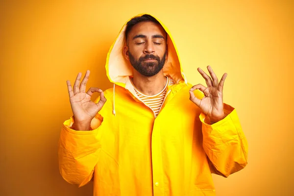 Young indian man wearing raincoat with hood standing over isolated yellow background relax and smiling with eyes closed doing meditation gesture with fingers. Yoga concept.