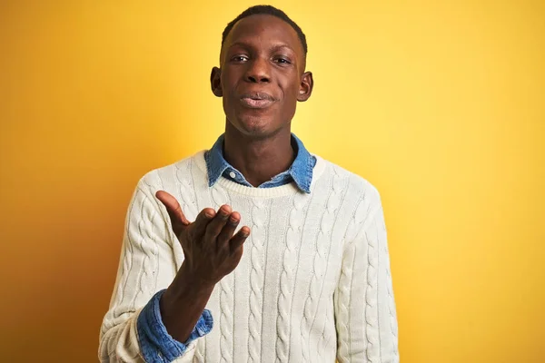 African american man wearing denim shirt and white sweater over isolated yellow background looking at the camera blowing a kiss with hand on air being lovely and sexy. Love expression.