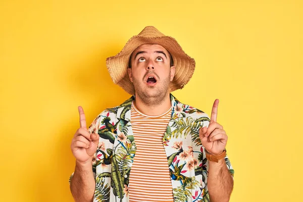 若い男オン休暇身に着けている夏のシャツと帽子上の隔離された黄色の背景驚きと驚きを見上げと指差す指と上げ腕 — ストック写真