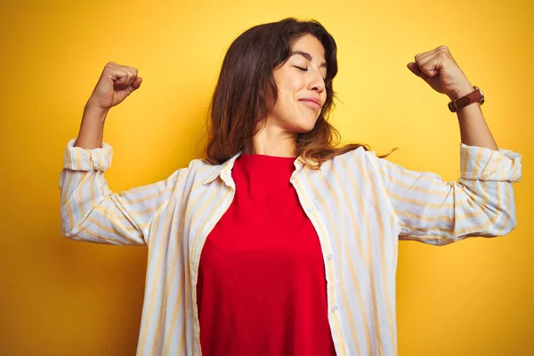 Young beautiful woman wearing red t-shirt and stripes shirt over yellow isolated background showing arms muscles smiling proud. Fitness concept.