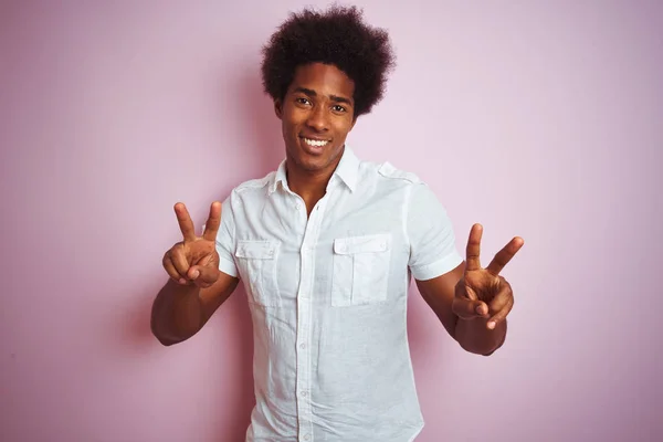 身穿白衬衫的非洲裔美国年轻人站在孤立的粉色背景上 微笑着看着摄像机 手指头在做胜利的标志 第二点 — 图库照片