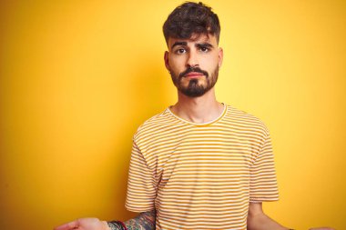 Çizgili tişörtlü genç adam izole edilmiş sarı arka planda duruyor. Hiçbir fikri yok ve kolları açık, hiçbir fikri yok..