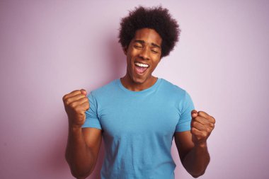 Afro saçlı, mavi tişörtlü, izole edilmiş pembe arka plan üzerinde mutlu ve heyecanlı bir şekilde duran, kollarını kaldırıp, gülümseyip başarı için bağıran Afro-Amerikan bir adam. Kutlama konsepti.
