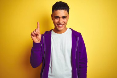 Mor kazak giyen genç Brezilyalı adam izole edilmiş sarı arka plan üzerinde duruyor ve bir numaralı parmağını gösterirken kendinden emin ve mutlu gülümsüyor..