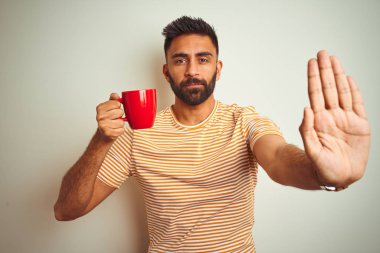 Genç Hintli bir adam kırmızı kahve içiyor. İzole edilmiş beyaz arka planda duruyor. Açık elle dur işareti yapıyor.