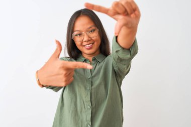 Yeşil gömlekli ve gözlüklü genç Çinli bir kadın izole edilmiş beyaz arka planda gülümsüyor ve mutlu yüzlü elleri ve parmakları olan bir çerçeve yapıyor. Yaratıcılık ve fotoğrafçılık kavramı.