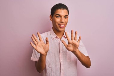 İzole edilmiş pembe arka plan üzerinde duran, rahat gömlekli genç yakışıklı Arap adam on numarayı gösterirken kendinden emin ve mutlu bir şekilde gülümsüyor..