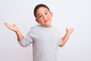 Gri günlük tişört giyen güzel çocuk izole edilmiş beyaz arka plan üzerinde duruyor. Kolları ve elleri havada, kafası karışık bir yüz ifadesi var. Şüphe kavramı.