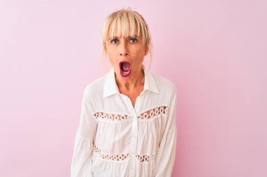 Beyaz gömlekli orta yaşlı bir kadın pembe arka planda dikiliyor. Şaşırtıcı bir ifade, korku ve heyecan dolu bir yüz ifadesi var..