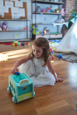 Anaokulunda bir sürü oyuncağın etrafında arabayla oynayan sevimli sarışın çocuk.