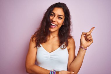 İzole edilmiş pembe arka planda duran beyaz tişörtlü genç ve güzel bir kadın. Yüzünde kocaman bir gülümseme var. Elleri ve parmakları kameraya bakıyor..