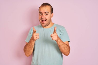 İzole edilmiş pembe arka planda duran mavi tişörtlü genç adam mutlu ve komik yüzlü parmaklarını kameraya doğrultuyor. İyi enerji ve titreşimler.