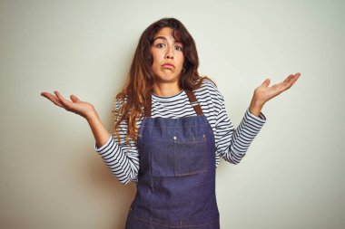 Önlük giyen genç, güzel bir şef kadın izole edilmiş beyaz arka planda yemek pişiriyor. Kolları ve elleri havada, kafası karışmış bir ifade takınıyor. Şüphe kavramı.