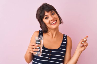 Genç ve güzel bir kadın pembe arka planda duran bir bardak suyu tutarken mutlu bir şekilde elini ve parmağını kenarı işaret ediyor.