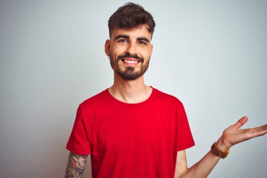 İzole edilmiş beyaz arka planda duran kırmızı tişörtlü genç adam gülümsüyor ve avuç içi kameraya bakıyor..