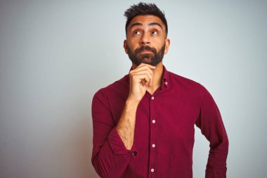 İzole edilmiş gri arka planda duran kırmızı şık tişörtlü genç Hintli adam elleri çenesinde soru sorarken düşünceli bir ifade takınıyor. Düşünceli bir yüzle gülümsüyor. Şüphe kavramı.