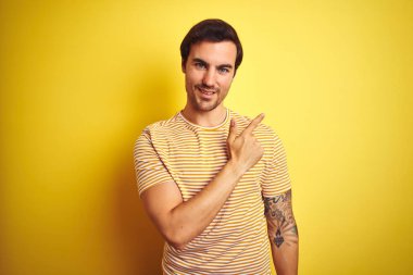 İzole edilmiş sarı arka plan üzerinde çizgili tişört giyen genç yakışıklı bir adam. Yüzünde mutlu ve doğal bir ifadeyle, elleri ve parmaklarıyla yüzünü gösteren bir gülümseme.