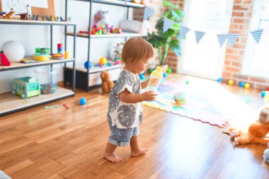 Anaokulunda bir sürü oyuncağın etrafında beslenme şişesi tutan sevimli çocuk.