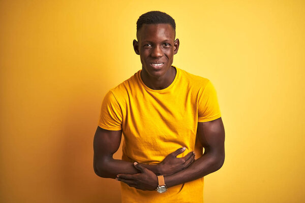 Young african american man wearing casual t-shirt standing over isolated yellow background with hand on stomach because indigestion, painful illness feeling unwell. Ache concept.
