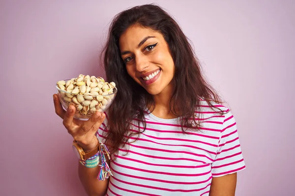 Young beautiful woman holding bowl with pistachios over isolated pink background with a happy face standing and smiling with a confident smile showing teeth