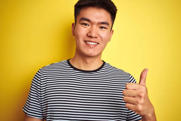 若いアジア系中国人男性を着てストライプTシャツ上に立って孤立した黄色の背景幸せな大きな笑顔でOk Signをやって 親指を指で開く 優れたサイン — ストック写真