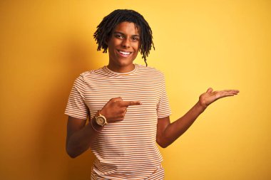 Sarı arka plan üzerinde duran çizgili t-shirt giyen Afro adam kameraya hayret ediyor ve gülümsüyor..