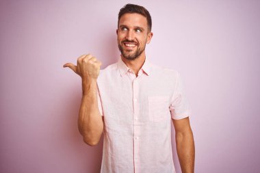 Pembe izole edilmiş arka planda zarif bir yaz gömleği giyen yakışıklı genç adam gülümsüyor ve başparmağıyla yüzünü yukarı kaldırıyor..