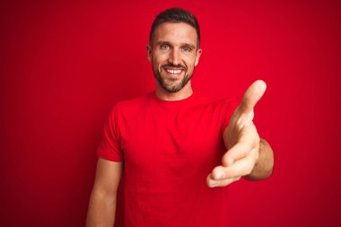 Kırmızı izole edilmiş arka plan üzerine gündelik tişört giyen yakışıklı genç adam dostça bir selamlama ve hoş geldin hediyesi olarak el sıkışma sunuyor. Başarılı bir iş..