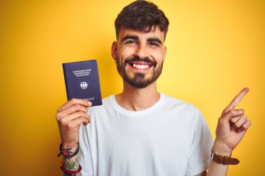 Alman pasaportu takan genç bir adam izole edilmiş sarı arka planda çok mutlu bir şekilde elini ve parmağını kenarı işaret ediyor.