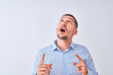 Genç yakışıklı iş adamı izole edilmiş arka planda duruyor. Şaşırmış ve şaşırmış bir şekilde yukarı bakıyor ve parmaklarıyla işaret ediyor ve kollarını kaldırıyor..
