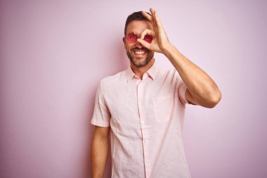 Zarif yaz gömleği ve güneş gözlüğü takan yakışıklı bir adam pembe soyutlanmış arka planda el hareketleriyle gülümsüyor ve mutlu yüzle parmaklarının arasından bakıyor..