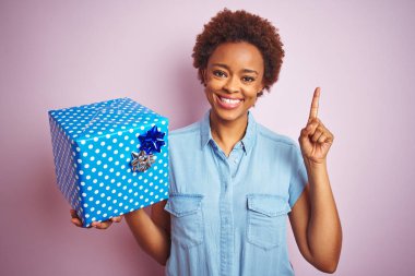 Doğumgünü hediyesini pembe izole edilmiş arka planda tutan Afrikalı Amerikalı kadın bir fikir ya da soru işareti ile şaşırdı.