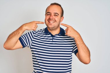 Sıradan çizgili polo giyen genç adam izole edilmiş beyaz arka planda gülümseyen ve parmaklarıyla ve ağzıyla işaret eden. Diş sağlığı konsepti.
