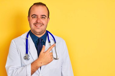Paltolu ve steteskoplu genç bir doktor, izole edilmiş sarı arka planda neşeli, yüzünde mutlu ve doğal bir ifadeyle el ve parmak ile gülümseyen bir gülümsemeyle dikiliyor.
