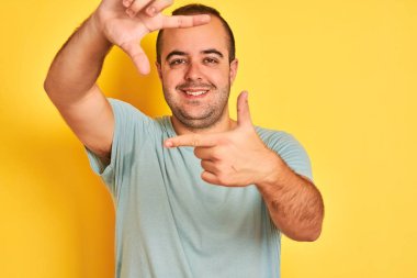 Yeşil günlük tişört giyen genç adam izole edilmiş sarı arka planda duruyor gülümsüyor ve mutlu yüzlü elleri ve parmakları olan bir çerçeve yapıyor. Yaratıcılık ve fotoğrafçılık kavramı.