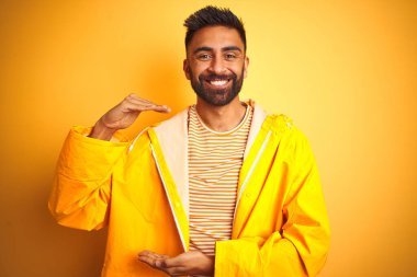 Yağmurluk giyen genç Hintli adam izole edilmiş sarı arka planda duruyor. Ellerinde büyük ve büyük boy işareti, ölçü sembolü var. Kameraya bakıp gülümsüyor. Ölçüm konsepti.