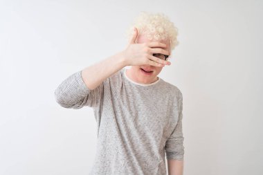 Genç, sarışın, çizgili tişört ve gözlüklü bir adam izole edilmiş beyaz arka plan üzerinde şok edici bir şekilde yüzünü ve gözlerini kapayarak utangaç bir ifadeyle parmaklarının arasından bakıyor..