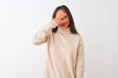 Balıkçı yaka kazak ve gözlük takan genç Çinli bir kadın izole edilmiş beyaz arka planda şok olmuş bir şekilde yüzünü ve gözlerini kapayarak utanmış bir ifadeyle parmaklarının arasından bakıyor..