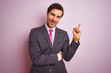 Takım elbiseli ve kravatlı genç yakışıklı bir işadamı pembe arka planda duruyor. Yüzünde kocaman bir gülümseme, eli ve parmağı kameraya bakıyor..
