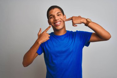 İzole edilmiş beyaz arka planda duran mavi tişörtlü genç, yakışıklı Arap adam gülümsüyor ve parmaklarıyla ve ağzıyla işaret ediyor. Diş sağlığı konsepti.