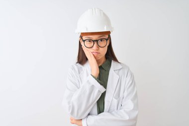 Genç Çinli mühendis kadın, izole edilmiş beyaz arka planda ceket kaskı gözlüğü takıyor ve kollarını kavuşturarak depresyondan sıkılmış ve yorgun görünüyor..