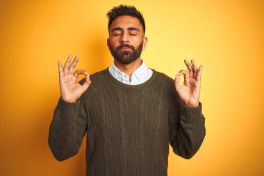 Yeşil kazak ve gömlek giyen genç Hintli adam izole edilmiş sarı arka planda duruyor rahatla ve gözleri kapalı gülümse parmaklarıyla meditasyon hareketi yapıyor. Yoga konsepti.