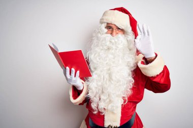 Orta yaşlı bir adam Noel Baba kostümü giyip izole edilmiş beyaz arka planda kitap okuyor gülümsüyor ve el ve parmaklarıyla 