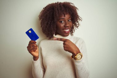 Kredi kartı tutan Afrikalı genç bir kadın izole edilmiş beyaz arka planda duruyor. El ve parmağıyla işaret etmekten çok mutlu.
