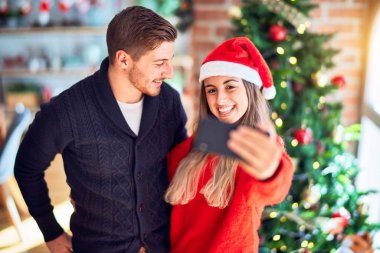Mutlu ve kendinden emin gülümseyen genç bir çift. Ayakta durmak ve sarılmak, evdeki Noel ağacının etrafında kamerayla selfie çektirir.