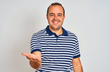 Sıradan çizgili polo giyen genç adam izole edilmiş beyaz arka planda durup neşeli gülümseyerek avuç içi yardım ve kabullenme teklif ediyor..