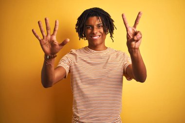 Rastalı tişörtlü Afro adam izole edilmiş sarı arka planda duruyor ve yedinci parmaklarını gösterirken kendinden emin ve mutlu bir şekilde gülümsüyor..