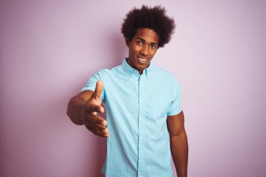Afro saçlı, mavi tişörtlü genç bir adam izole edilmiş pembe arka planda durup dostça gülümseyerek selamlama ve hoş geldin diyor. Başarılı bir iş..