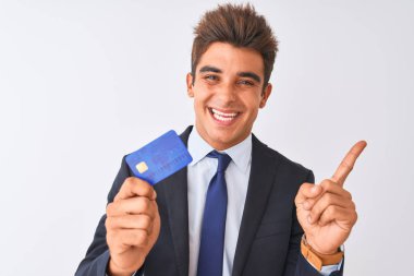 Soyutlanmış beyaz arka planda kredi kartı tutan takım elbiseli genç ve yakışıklı bir işadamı elini ve parmağını kenarı işaret ederek çok mutlu olur.